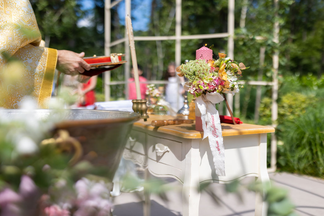 Cadru surprins de fotograf la botez cu un gheridon de restaurant improvizat de preot in gradina restaurantului ca sa tina loc de analoghion, cu lumanarea de botez, Sfanta Evanghelie si Sfanta Cruce.