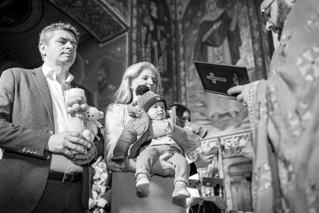 nasii cu bebelusul in brate in fata preotului care citeste din evanghelie, surprinsi de fotograf la botez