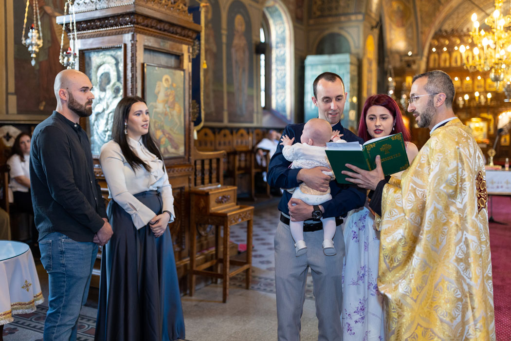 Fotograful de botez surprinde nasii cu pruncul in brate, alaturi de parinti, citind Crezul din evenghelia tinuta de preot.